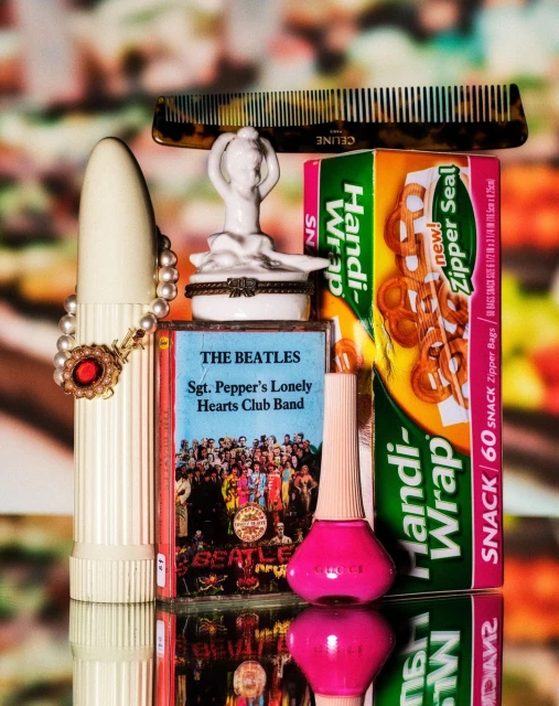 Still life studio shot of The Beatles album, comb, nail polish, and figurine on a reflective surface with a blurred background.