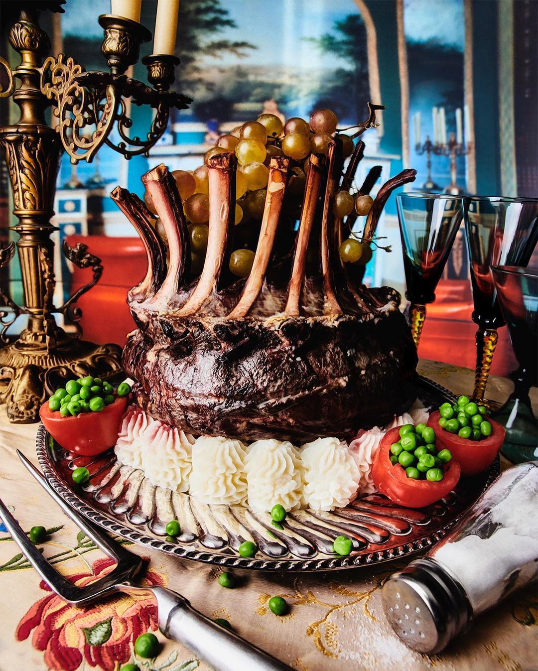 Elaborate rack of lamb dish with grapes, peas, and decorative silverware on a floral tablecloth, set against an ornate backdrop.