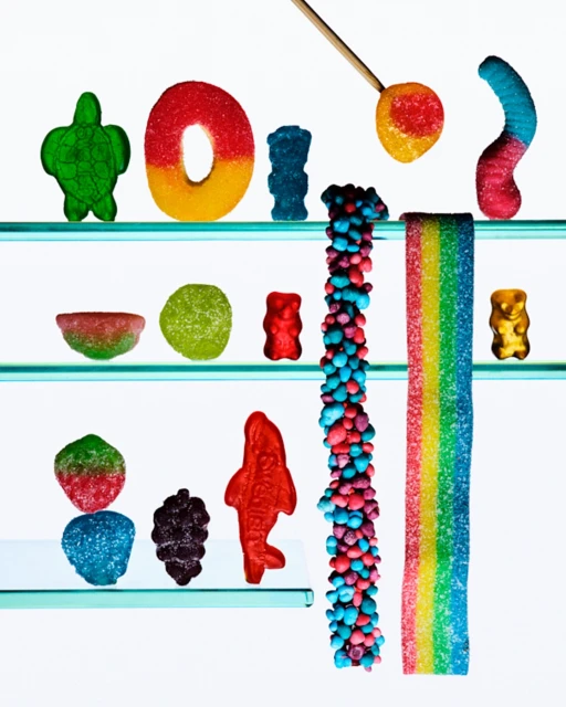 Assortment of colorful gummy candies on clear glass shelves, including worms, bears, and fish, against a bright white background.