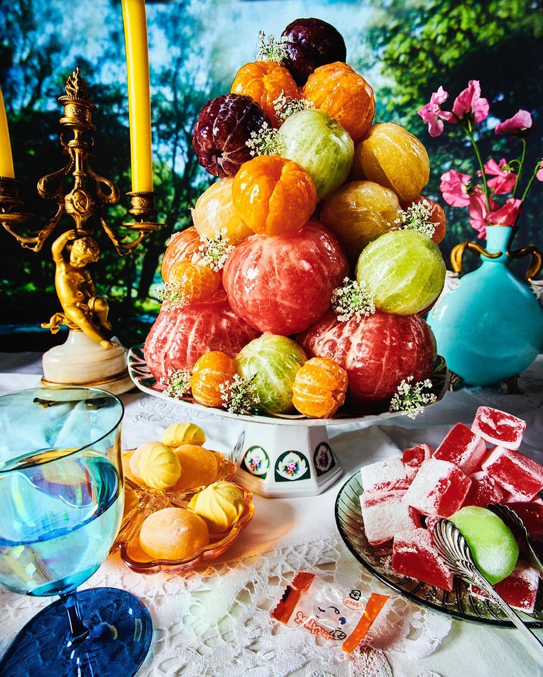 Still life of citrus fruit pyramid surrounded by hard candies, glass dishware, ornate candle holder, and decorative table cloth.