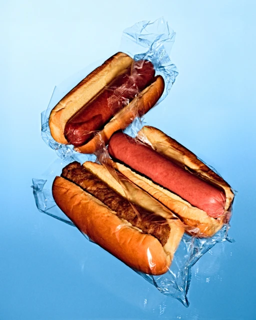 Overhead view of three hot dogs in buns, partially wrapped in clear plastic, displayed against a smooth, light blue backdrop.