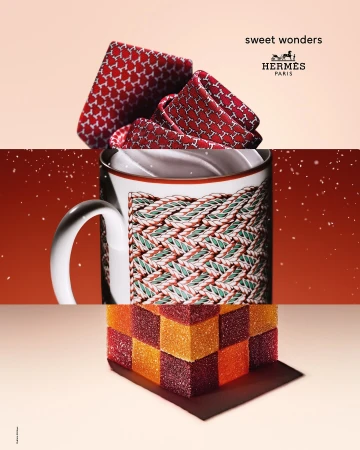 Hermes branded still life of folded scarf, mug, and cube of orange and red marmalade squares on a gradient pink background.