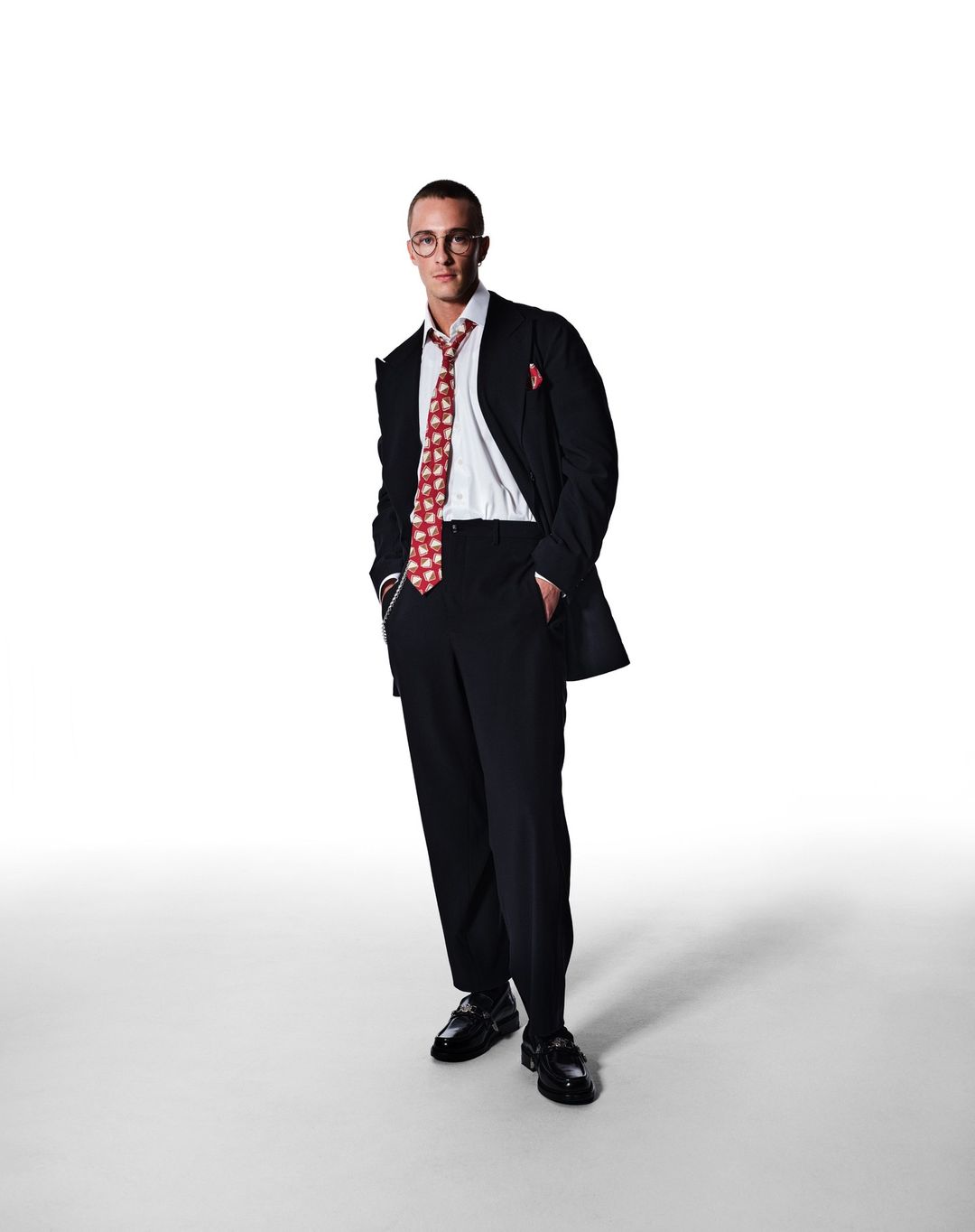 Full-length portrait of a man in a black suit and patterned red tie against a white background, wearing glasses and polished shoes.