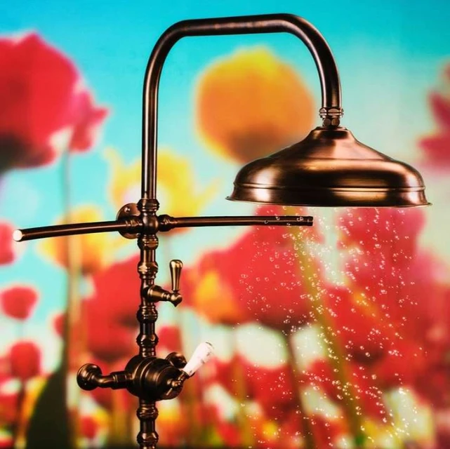 Close-up of a bronze shower fixture showing water droplets falling, set against a blurred background of red, orange, and blue flowers.