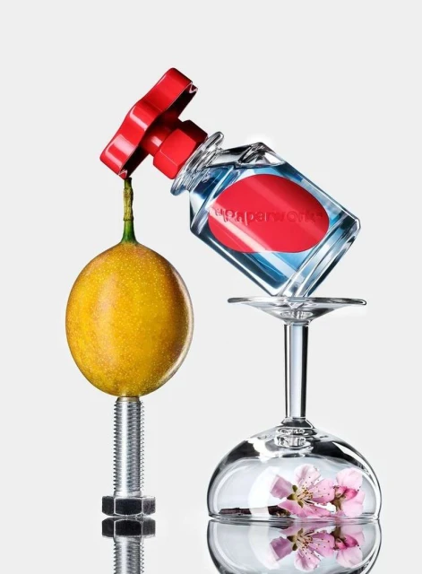 Still life composition of perfume bottle, yellow fruit secured by bolt, and wine glass with pink flowers on a white background.