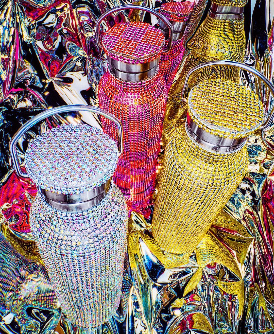 Several colorful and decorated water bottles with rhinestone details sit on a shiny and colorful reflective surface in a vibrant composition.
