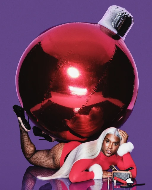 Adult poses indoors next to a large, red Christmas ball ornament, wearing a red, fur-trimmed bodysuit, fishnet tights, and black high heels.