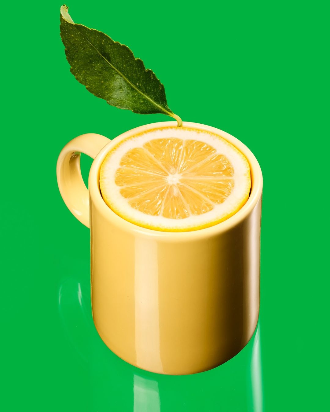 Close-up of a yellow mug filled with a lemon slice and leaf against a green background in studio lighting, showcasing a refreshing beverage.