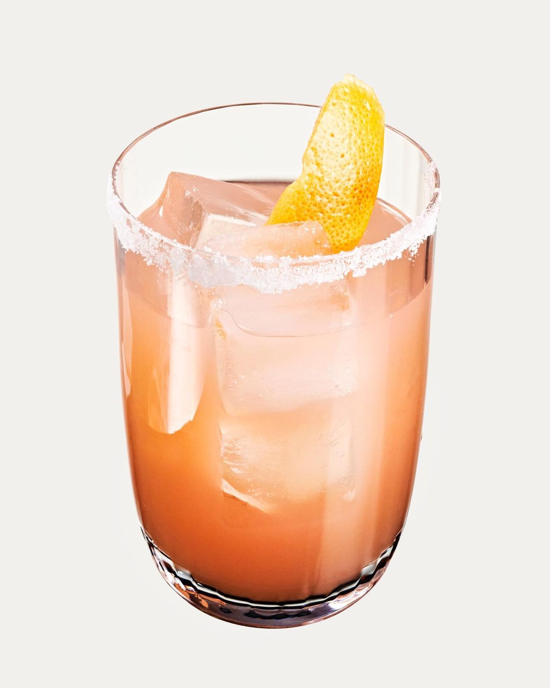 Close-up of a salty paloma cocktail in a clear glass with ice cubes and a citrus peel garnish against an off-white backdrop.