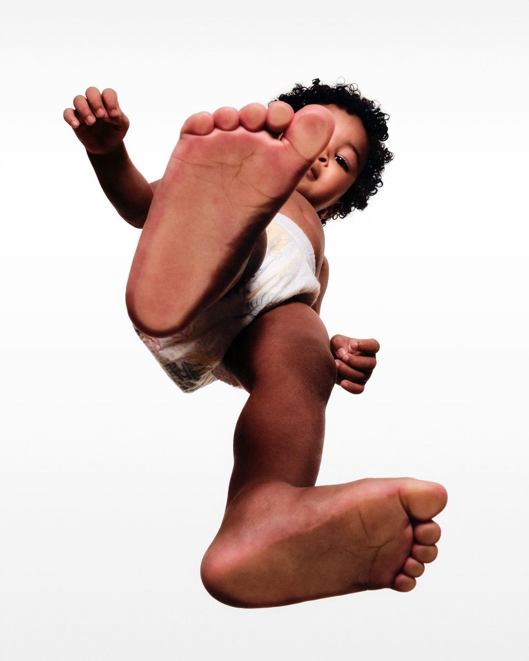 Low angle of a toddler in a diaper with curly black hair looking down, with one foot extended toward the viewer against a white background.