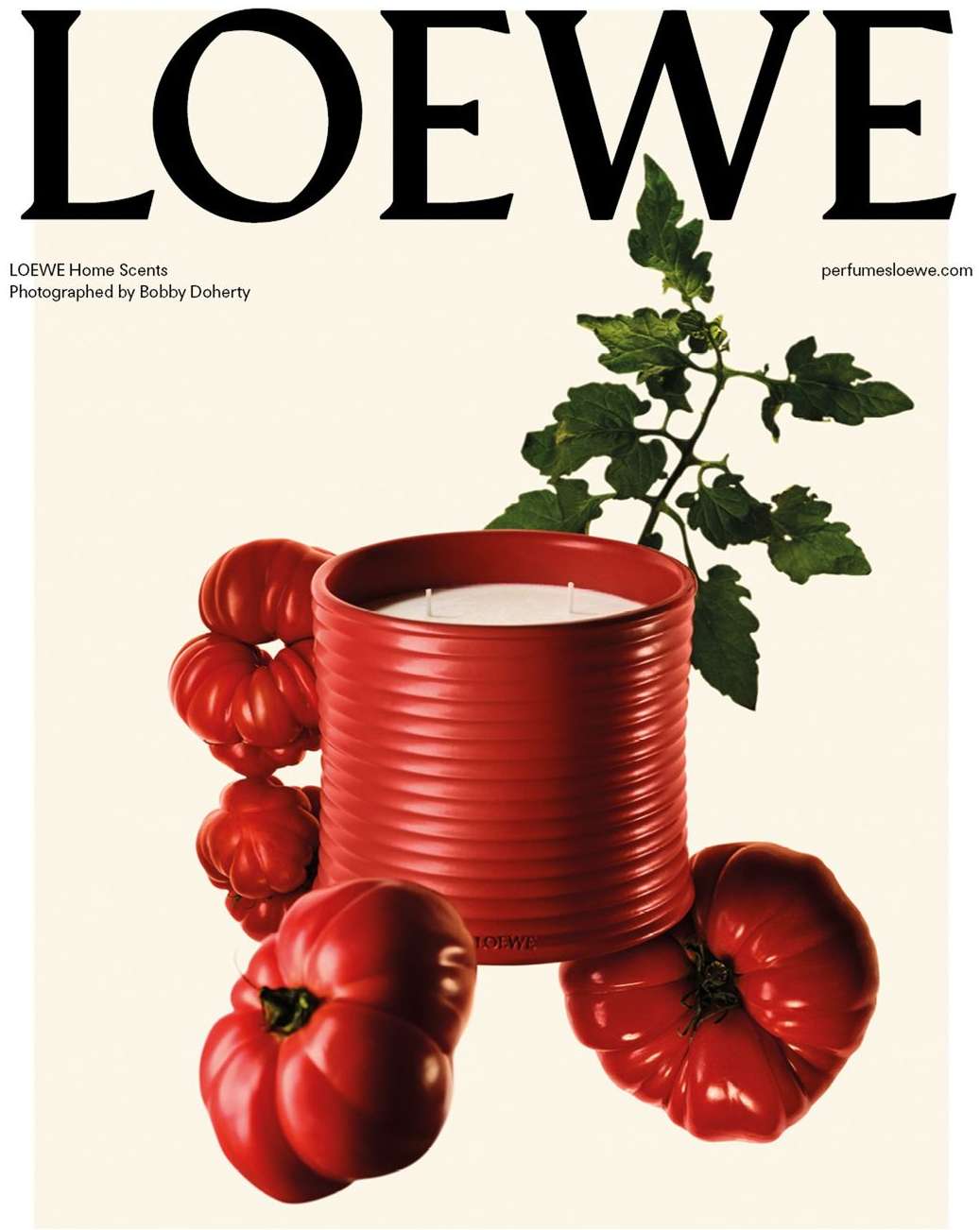 Loewe Home Scents ad featuring a red ribbed candle surrounded by red heirloom tomatoes and a tomato vine against a cream backdrop.