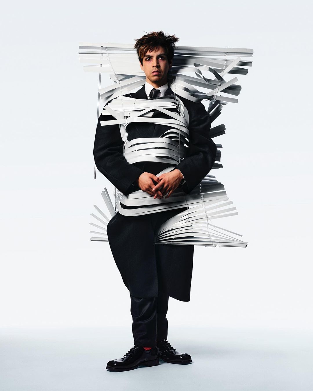 Young man stands in full suit wrapped in white window blinds against a bright white background in a studio setting for a fashion editorial.