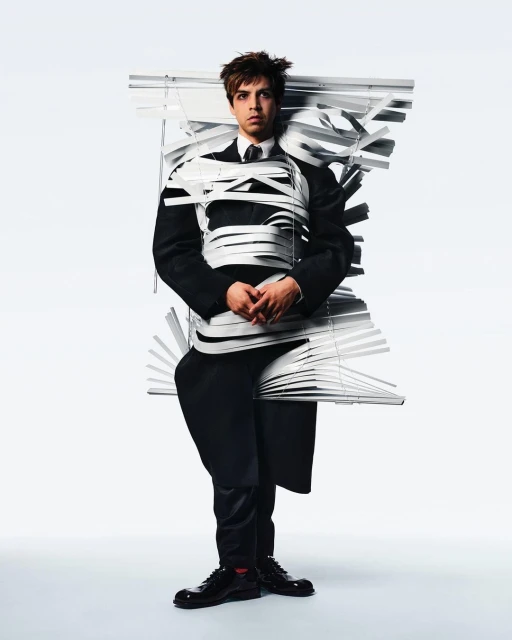 Young man stands in full suit wrapped in white window blinds against a bright white background in a studio setting for a fashion editorial.