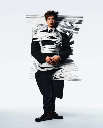 Young man stands in full suit wrapped in white window blinds against a bright white background in a studio setting for a fashion editorial.