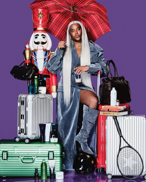 Person with long silver wig, wearing a blue velvet coat, standing among suitcases, a nutcracker, umbrella and various beauty products.