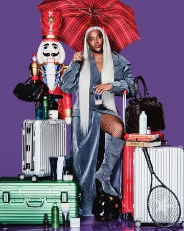 Person with long silver wig, wearing a blue velvet coat, standing among suitcases, a nutcracker, umbrella and various beauty products.