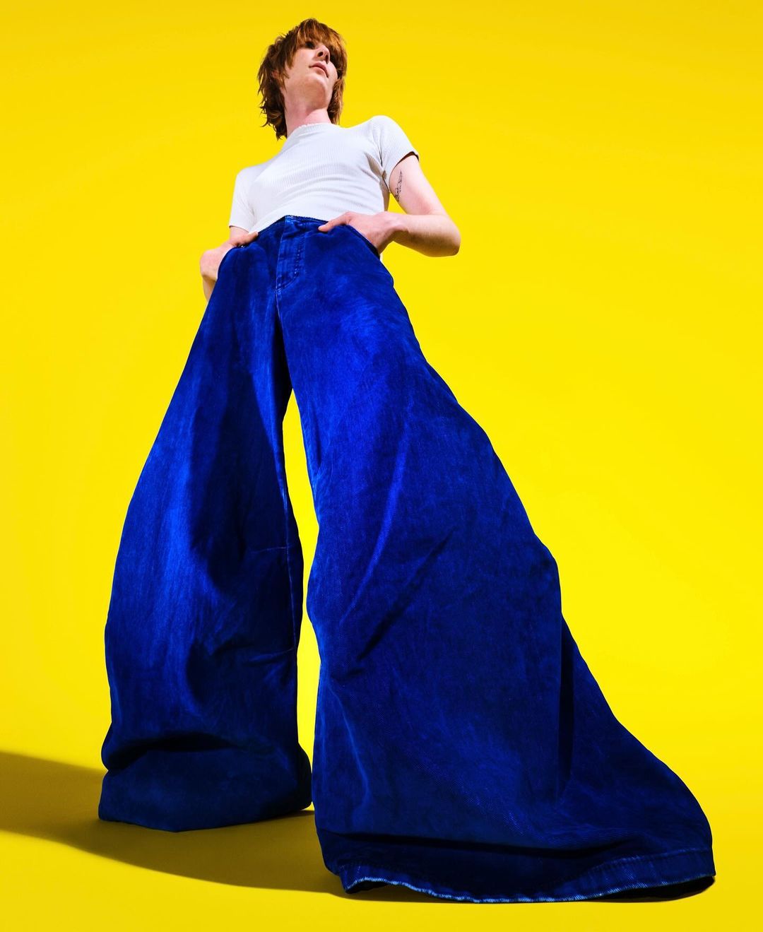 Person in a white t-shirt and oversized royal-blue bell-bottom jeans stands on a bright yellow background, hands in pockets.