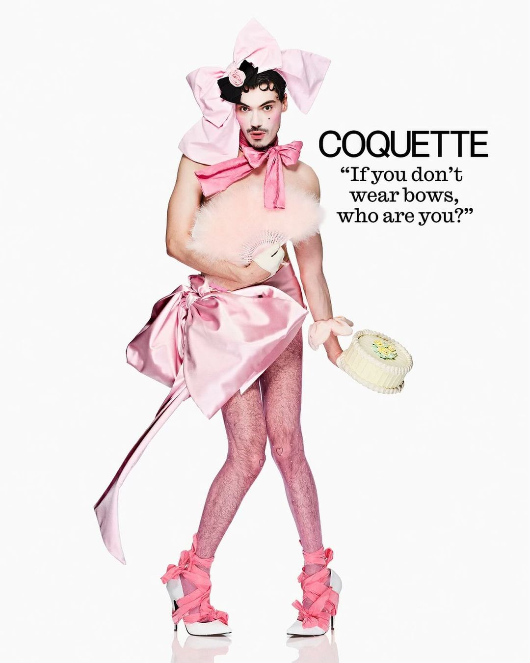 A person in pink drag attire poses in a full shot against a white backdrop, holding a feather fan and small cake.