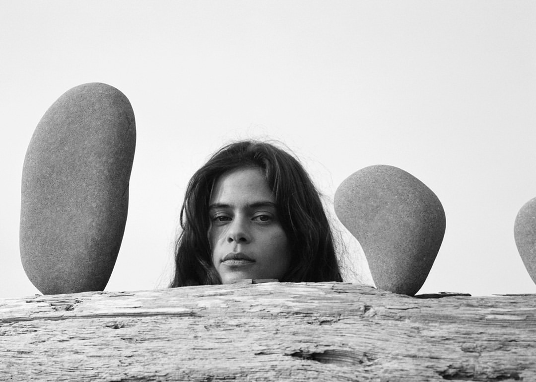 Black and white portrait of a woman's face between stones on weathered wood. The image is a simple composition with soft lighting.
