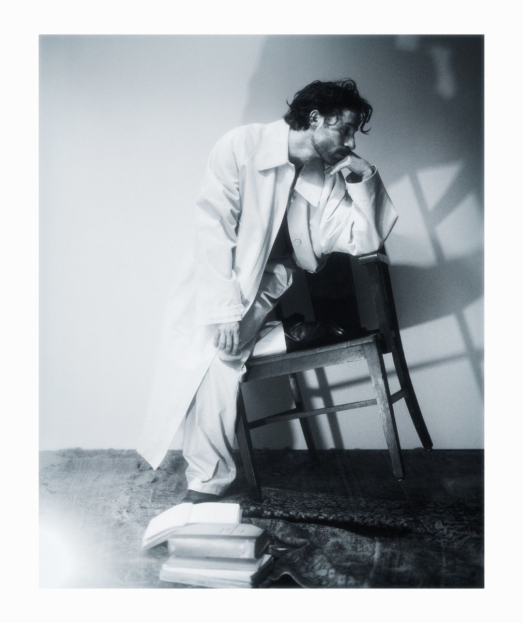 Man in white coat seated on a wooden chair next to stack of books against a wall with light and shadow play, fashion editorial.