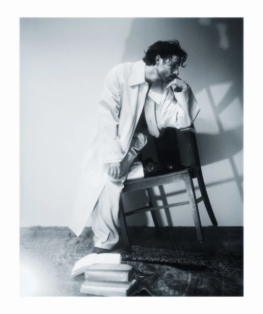 Man in white coat seated on a wooden chair next to stack of books against a wall with light and shadow play, fashion editorial.