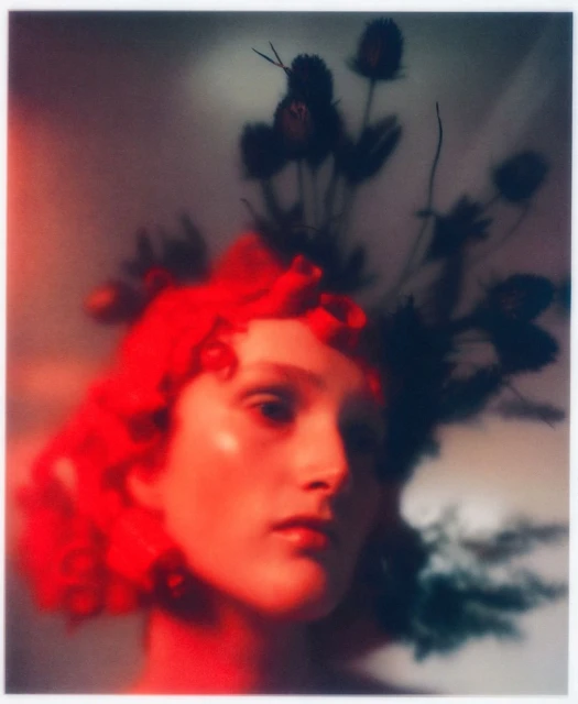 Portrait of a woman with bright red hair and headpiece against a soft gradient background with visible artistic blur and colour cast.
