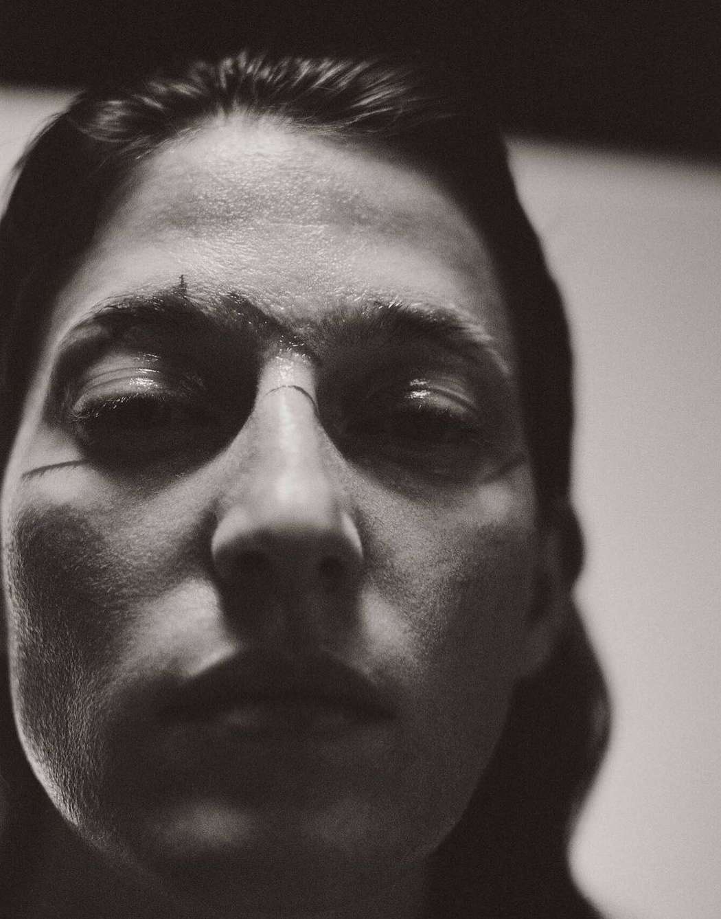 Close-up black and white portrait of a person with markings on their face looking directly at the camera with soft, diffused lighting.