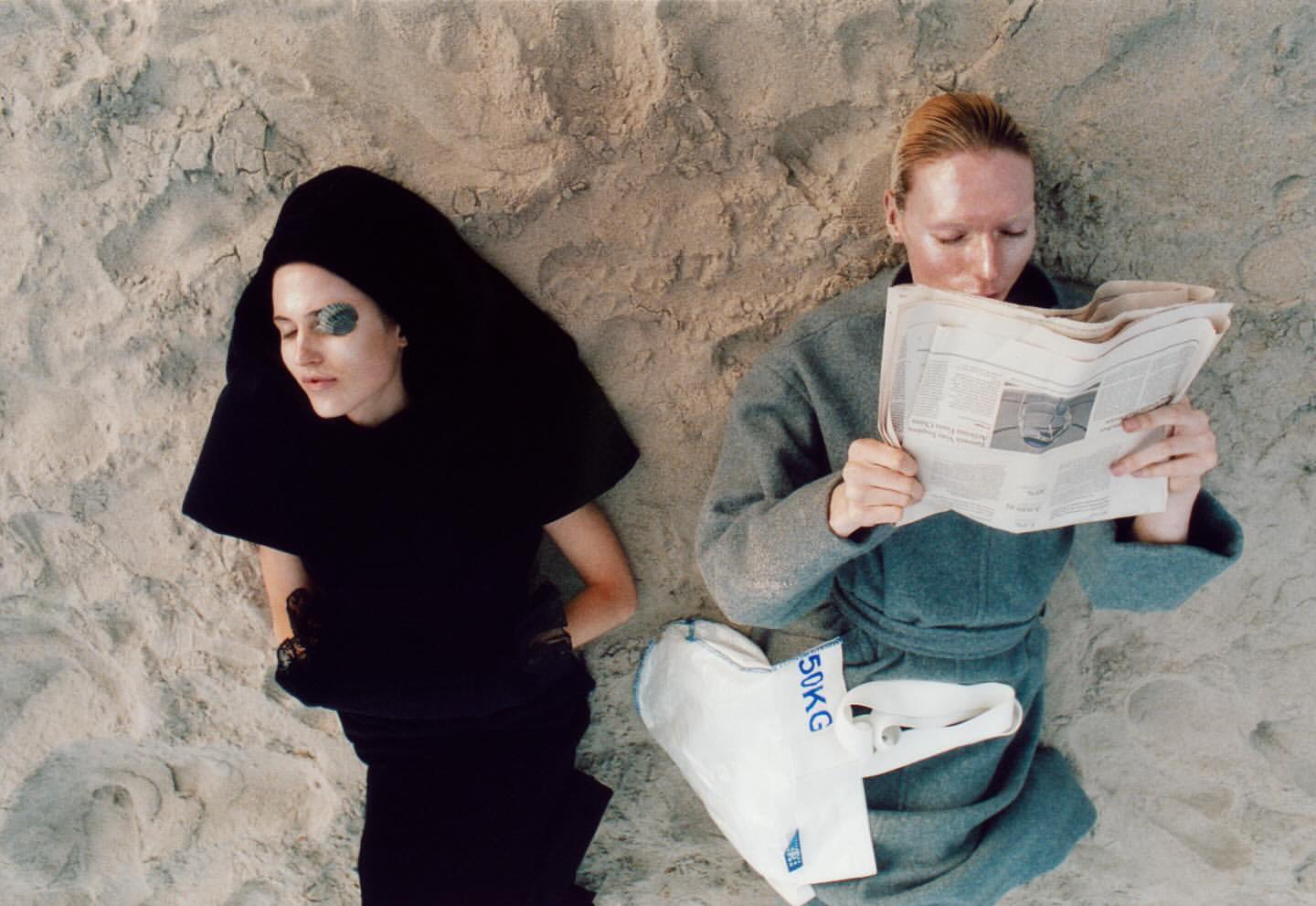 Two women lie on a sandy beach, one reads a newspaper while the other wears an eye patch, both dressed in fashion forward outfits.