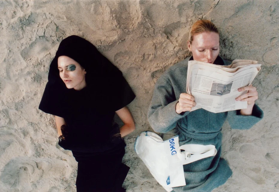 Two women lie on a sandy beach, one reads a newspaper while the other wears an eye patch, both dressed in fashion forward outfits.