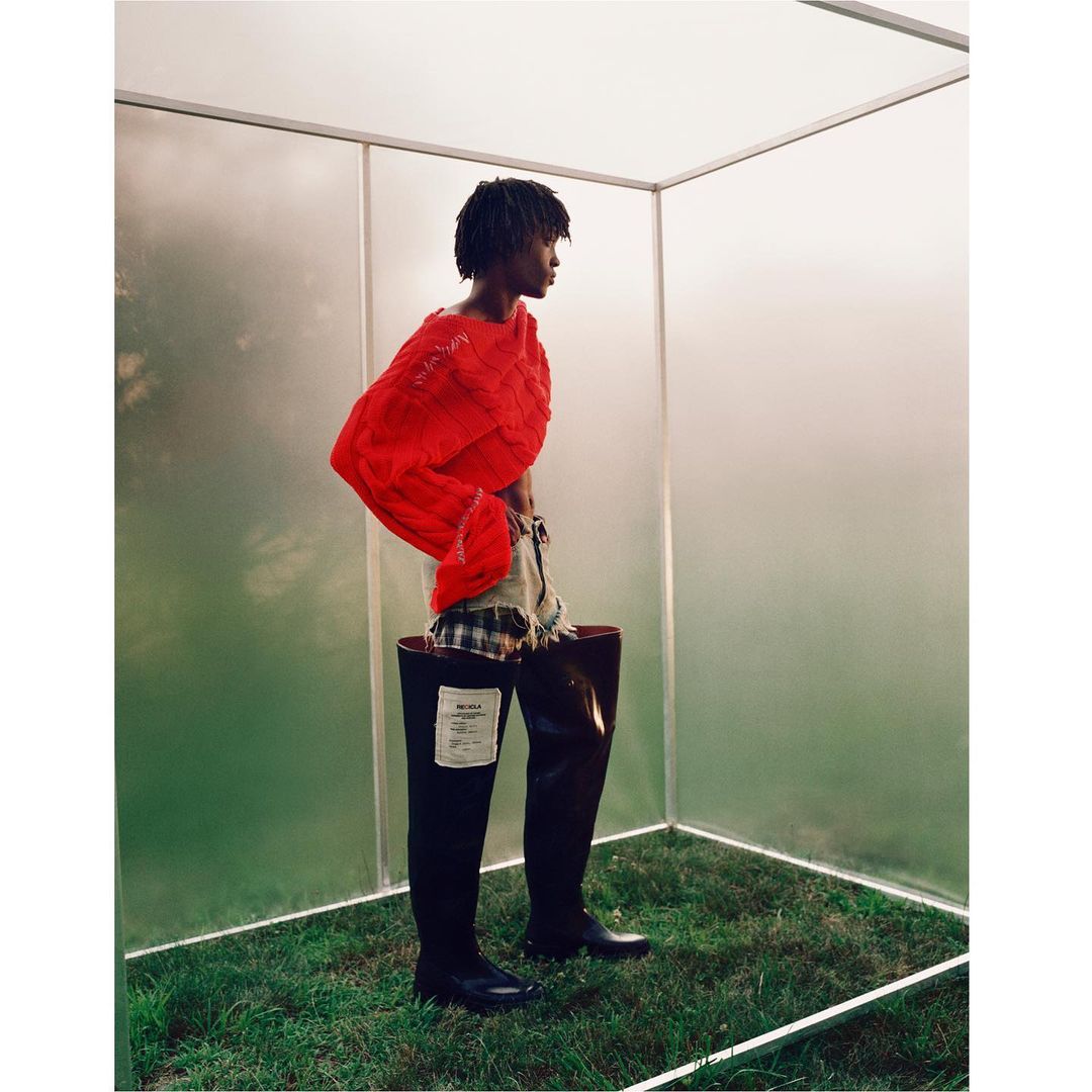 A person stands in oversized black boots inside a clear box on green grass. The model wears a red knit shirt and plaid shorts.