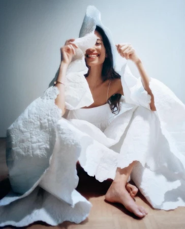 Smiling woman wrapped in a textured white quilt holding up a corner against a plain light wall