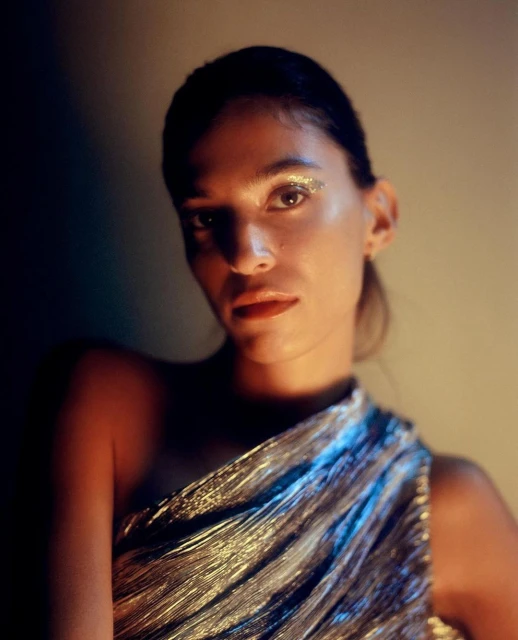 Portrait of a woman with glitter eyeshadow wearing a metallic silver one-shoulder dress against a dark background in soft, diffused light.