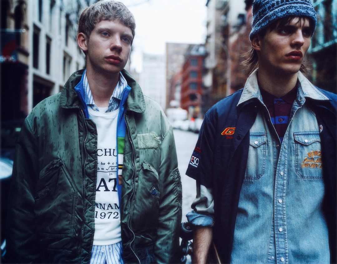 Two young adult male fashion models pose on a city street. One wears a green jacket, the other a denim shirt and blue hat.