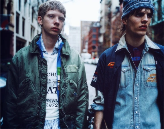 Two young adult male fashion models pose on a city street. One wears a green jacket, the other a denim shirt and blue hat.