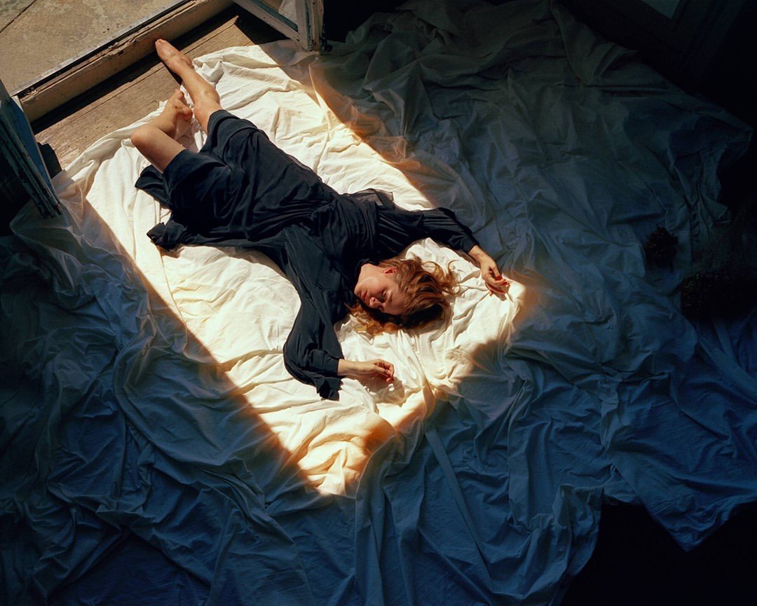 Woman in long dark dress laying on wrinkled white fabric in a sunlit room with open door, shot from overhead in landscape orientation.