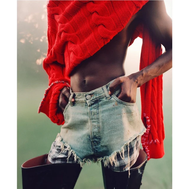 Fashionable person poses in cropped jean shorts, plaid undershorts, and a chunky red sweater, with black boots and an outdoor setting.