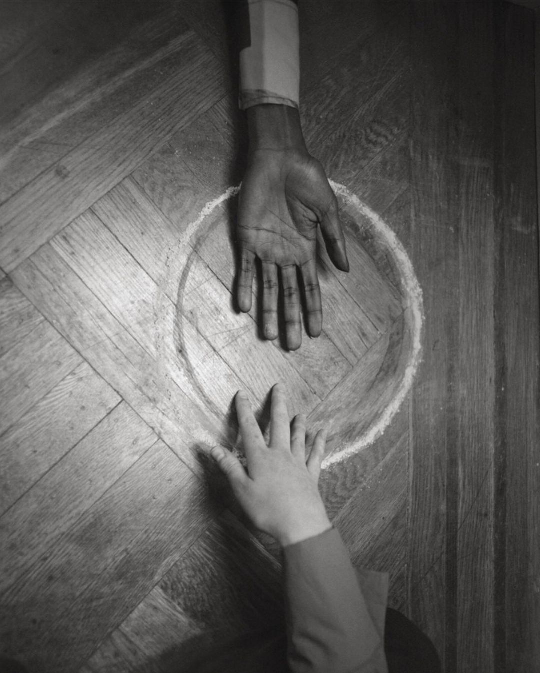 High-angle shot of two hands, one dark-skinned, one light-skinned, reaching toward each other inside a drawn chalk circle on a wood floor.