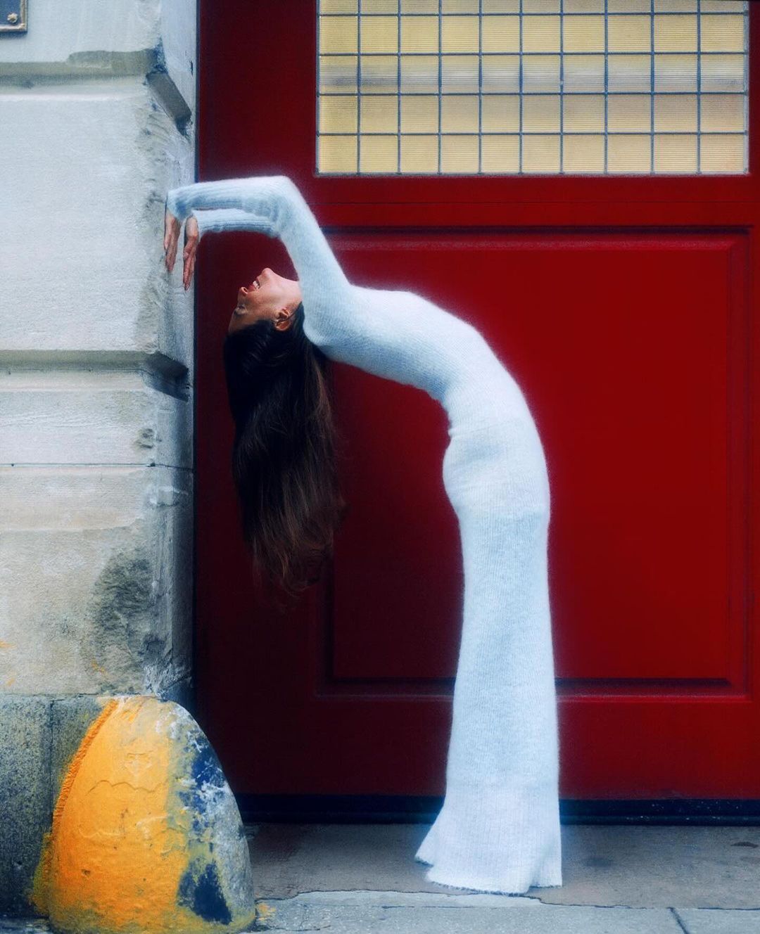 A slender female model in a white dress gracefully arches her back against a red door and weathered building facade in an urban setting.