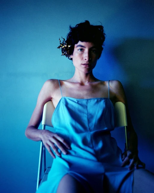 A young adult woman is sitting on a chair, looking at the camera, wearing a light blue dress, with a blue wall in the background.