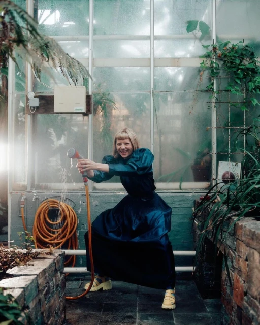 A smiling young woman in an emerald dress stands in a greenhouse, spraying water from a hose; plants and sunlight create a fresh, natural setting.