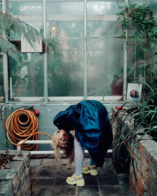 Adult female in blue satin dress bends over backward, showing blonde hair, pale tights, and yellow platform shoes in a greenhouse.
