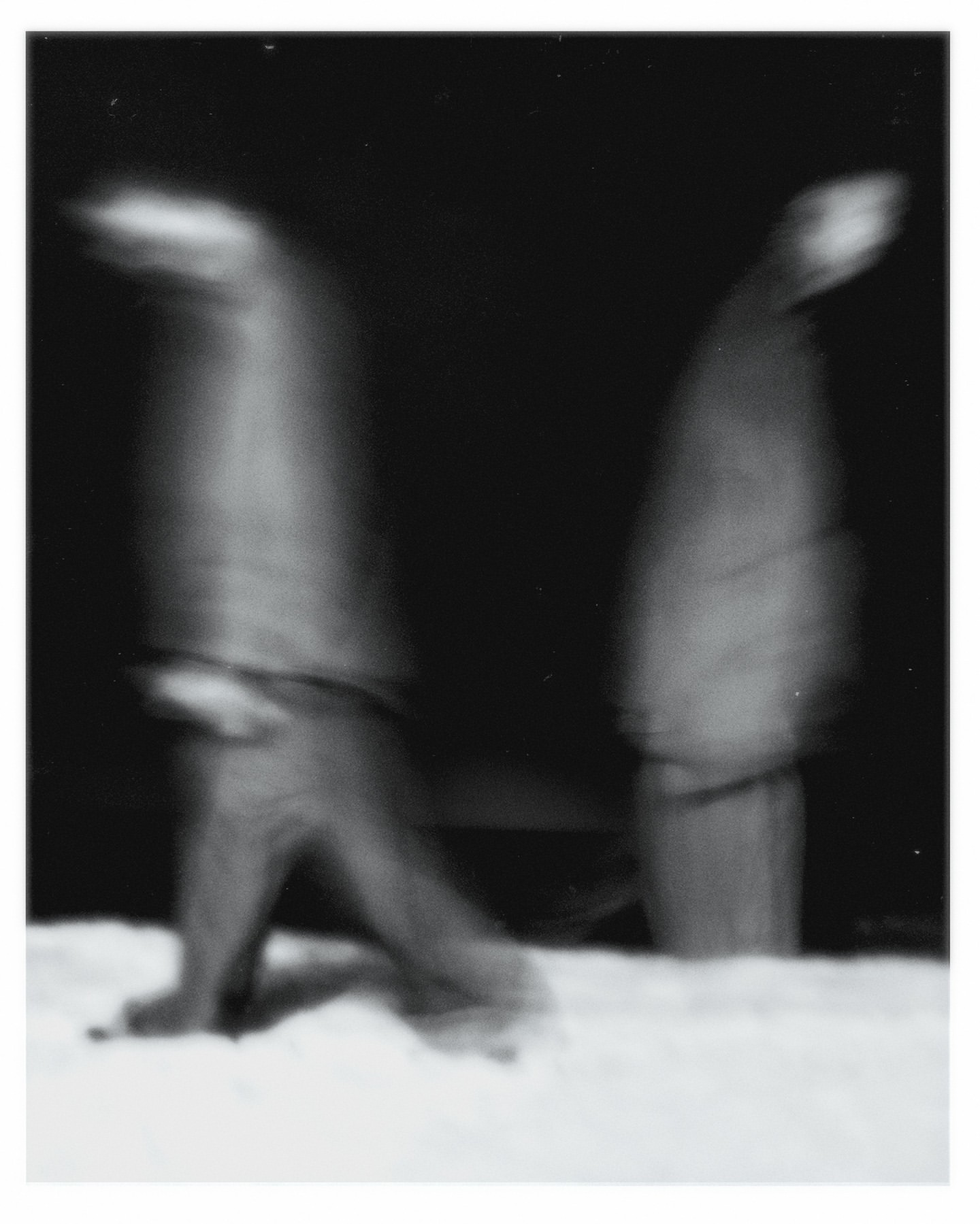 Monochrome long exposure shows two blurry figures walking on snow at night, creating a sense of movement and abstraction in a wintery scene.