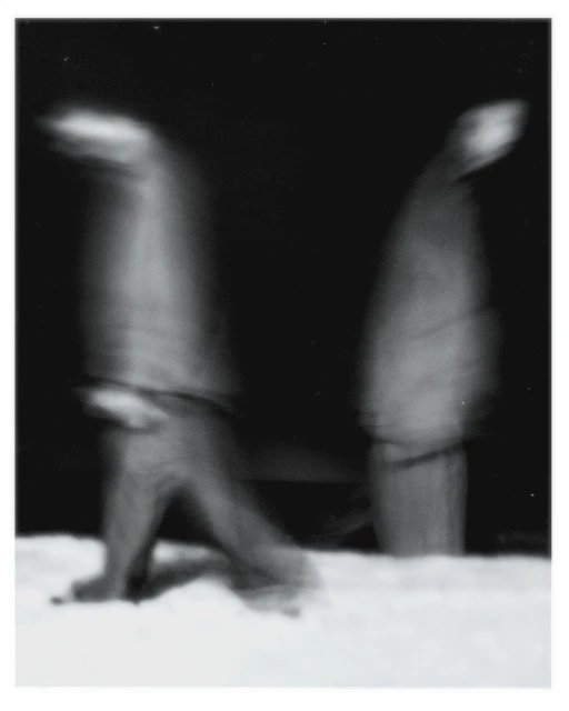 Monochrome long exposure shows two blurry figures walking on snow at night, creating a sense of movement and abstraction in a wintery scene.