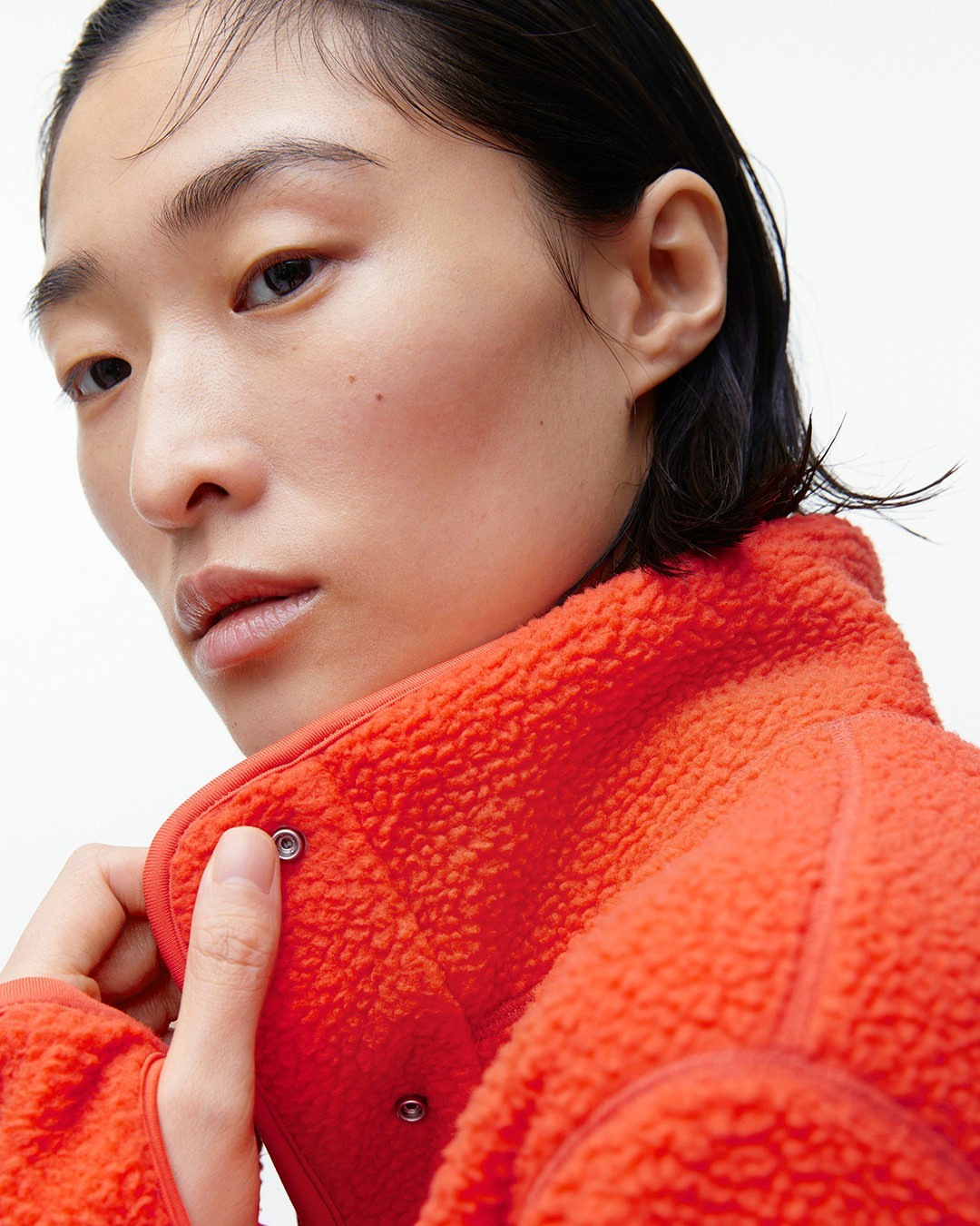Close-up of a young adult woman with fair skin wearing a bright orange fleece jacket, set against a stark white backdrop.
