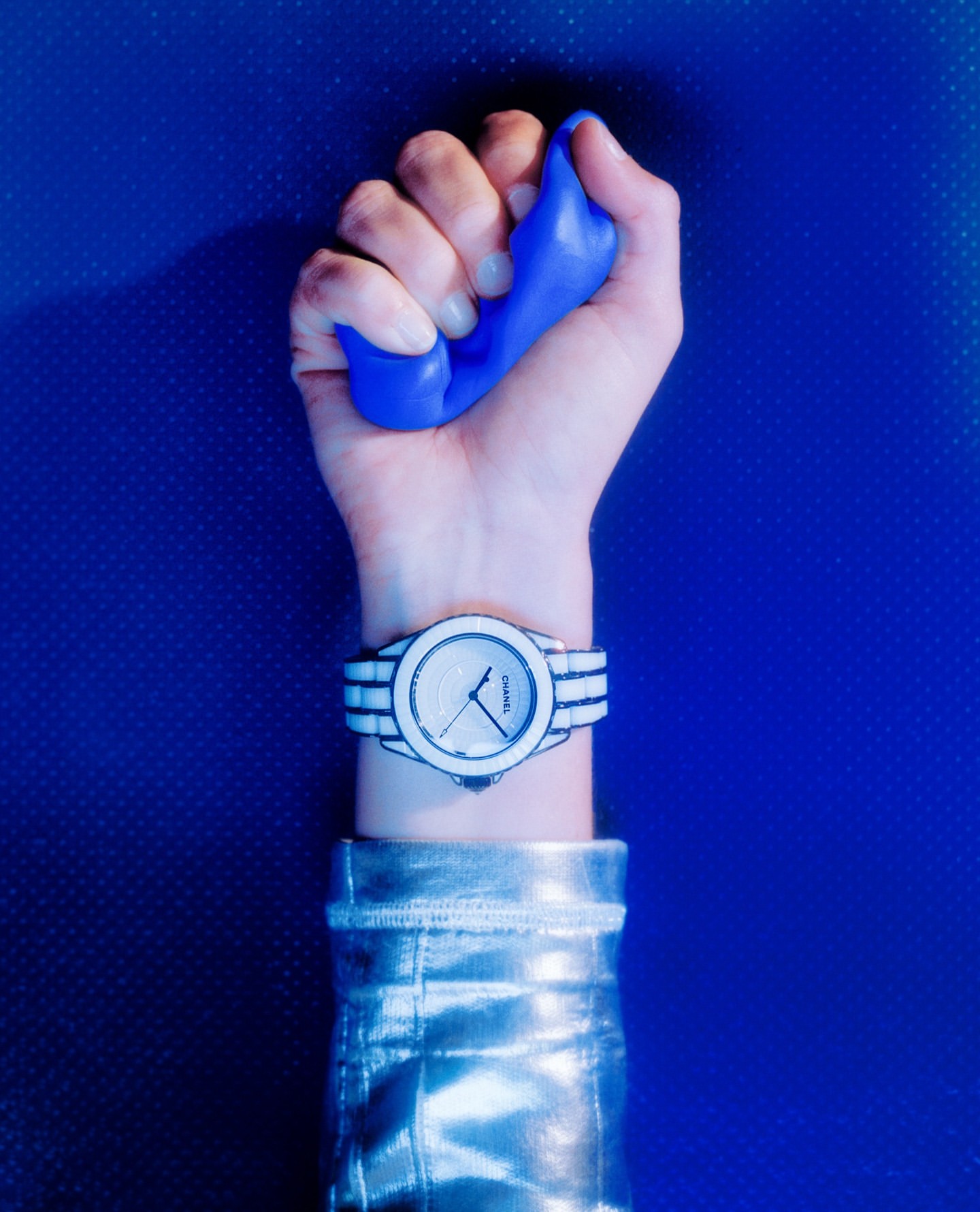 Woman's hand squeezing blue putty, wrist watch, and silver jacket sleeve against a blue background. Close-up product shot.