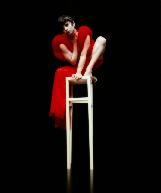 A slender individual perches on a white stool, draped in a vibrant red dress against a stark black background, illuminated by soft lighting.