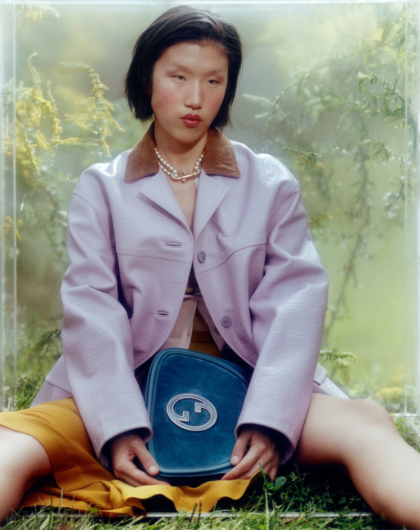 Asian model poses in a lilac jacket with a blue Gucci handbag in an outdoor setting with a light, diffused background.