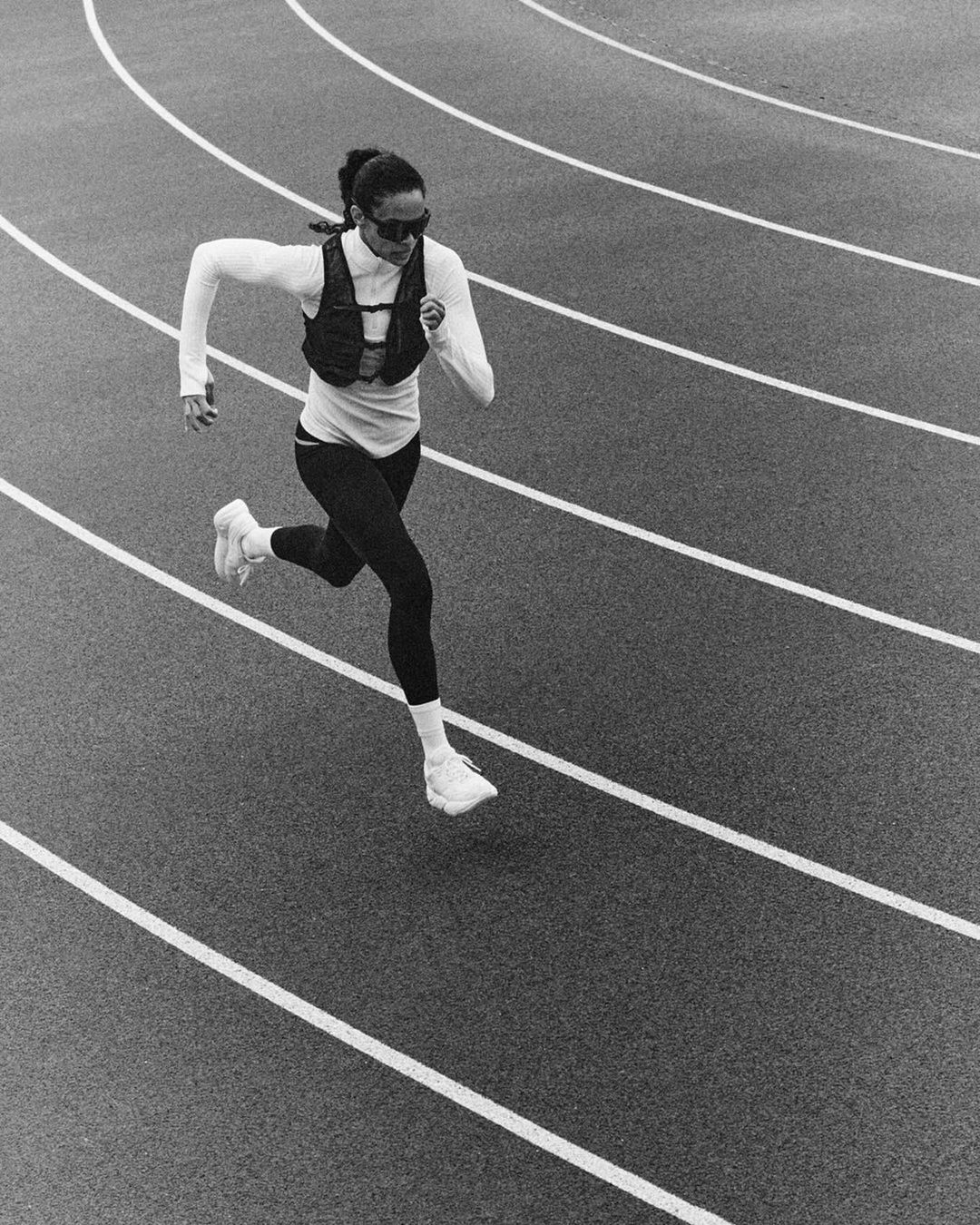 A woman runs on a track in monochrome, wearing sunglasses, a long-sleeved shirt, leggings, running shoes, and a vest with a water bottle.