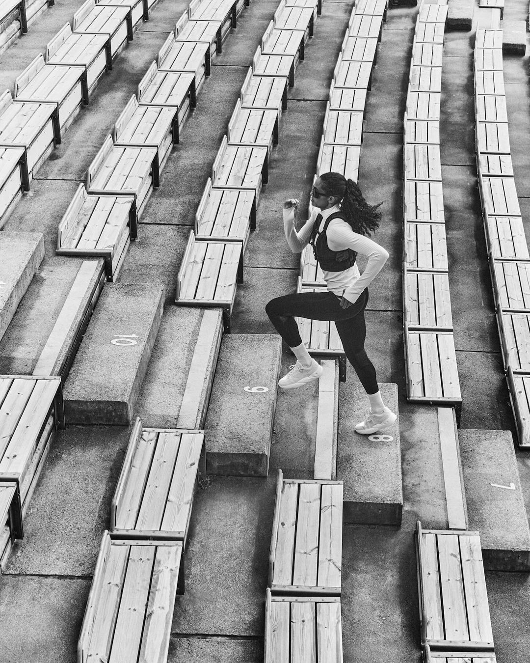 Woman in sportswear runs up stadium steps in black and white, showcasing fitness and determination in an urban setting.