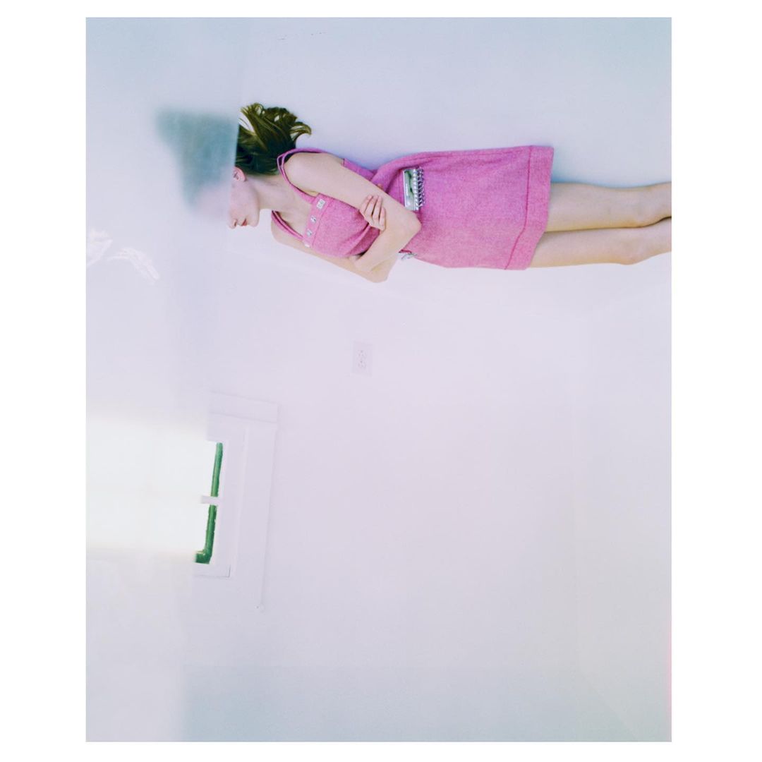 A woman lies on her side wearing a pink dress in a bright white room with a window. The image is high-key and in portrait orientation.